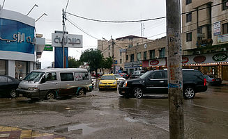 Eine Straße bei Regen: es hat sich eine große Pfütze gebildet, durch welche jetzt Autos, Busse und FußgängerInnen hindurch müssen. (Foto: EMS/Janke)